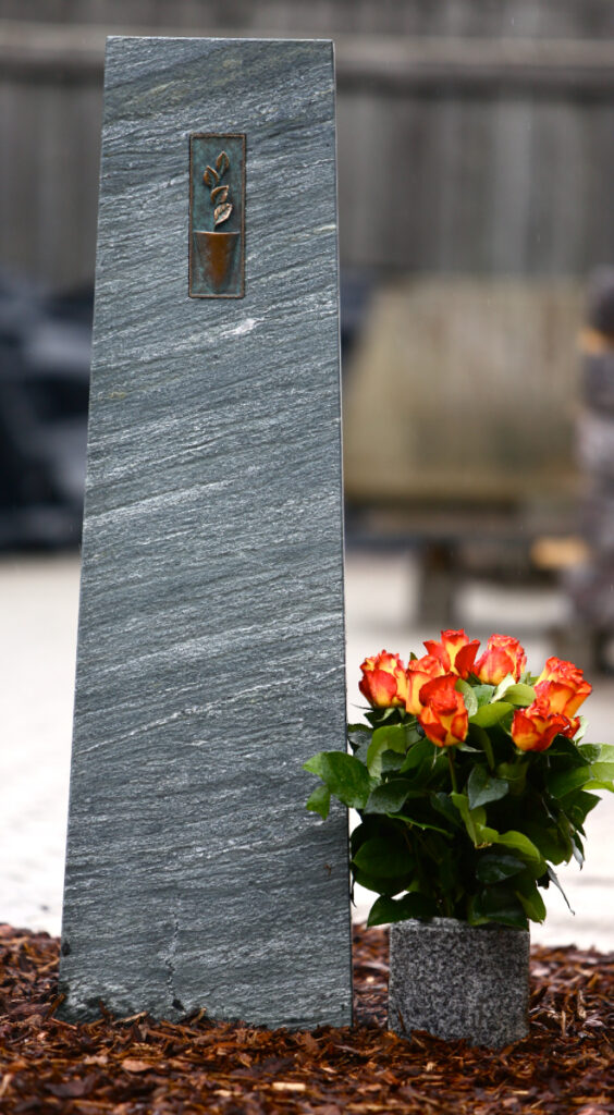 Elegant mindesten i grå natursten med bronzedekoration og orange roser.