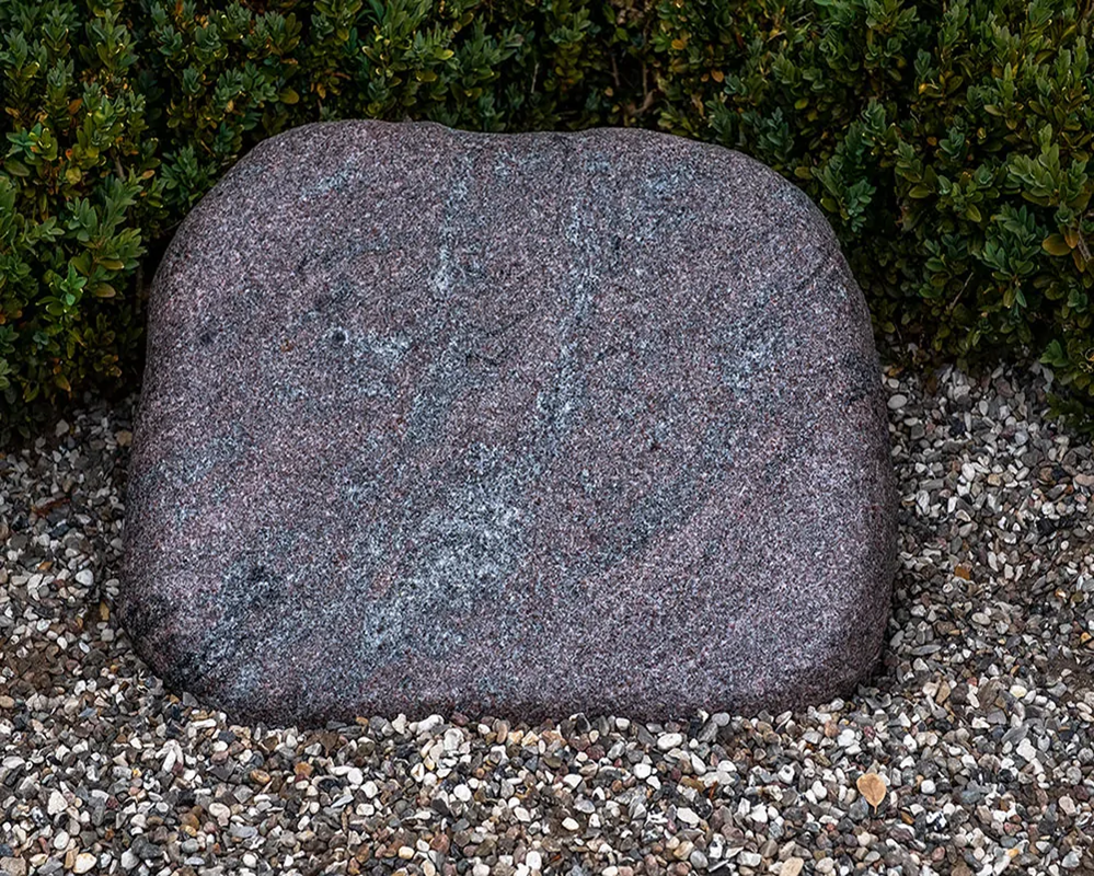 Skråtstillet plænesten i rå granit placeret i græsplæne på kirkegård.
